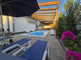 Un área de piscina con sillones de descanso y sombrillas, rodeada por una pérgola de madera y plantas en macetas.