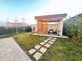 Un patio exterior moderno con una pérgola de madera, una parrilla de barbacoa y una zona de estar con mesa y sillas.