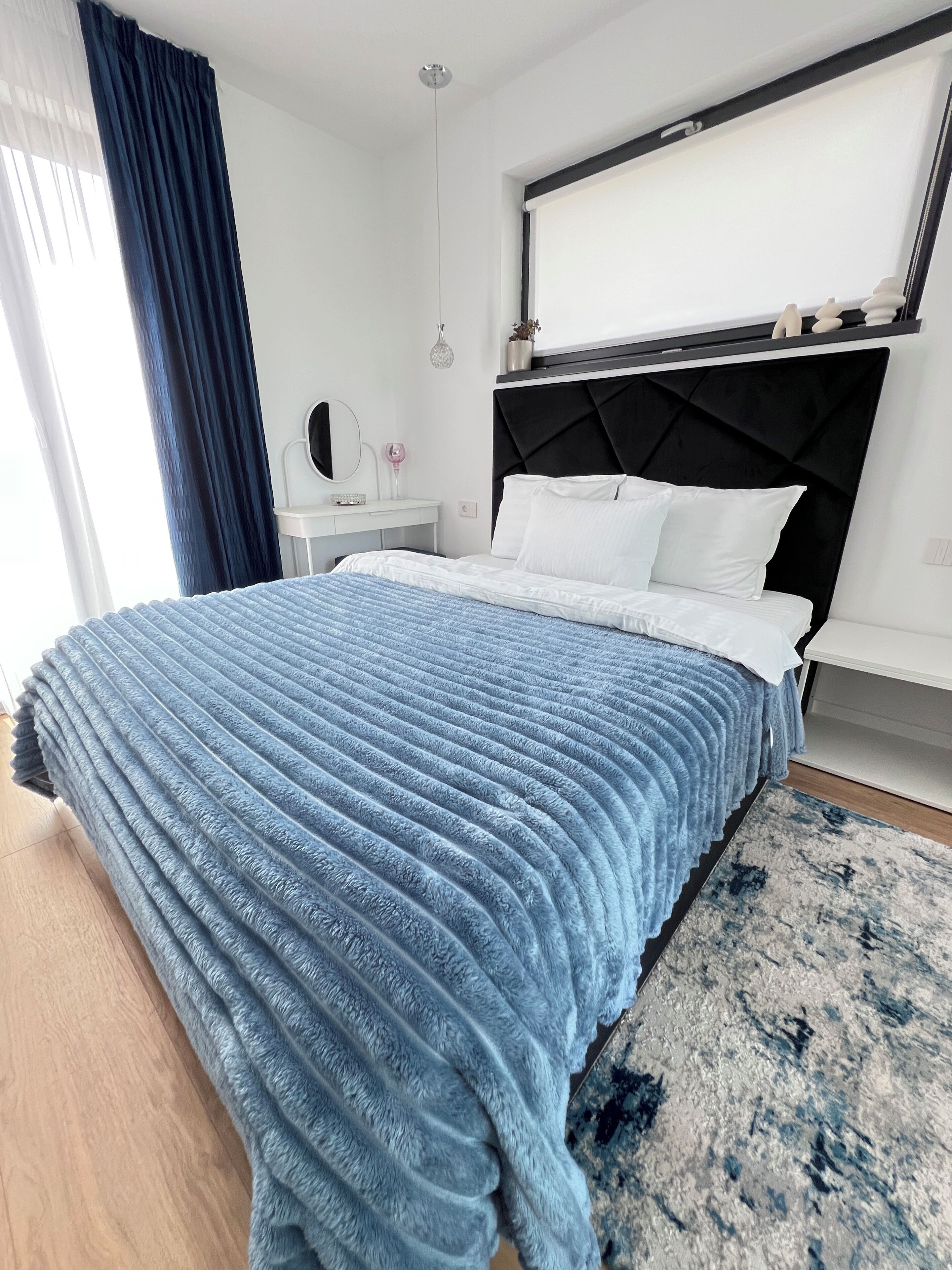 A modern bedroom with a large bed, a bedside table, a dresser, a mirror, and a window with curtains.