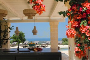 Un espace extérieur abrité avec vue sur la mer, une table garnie de fruits et d’un saladier, ainsi que des luminaires suspendus.