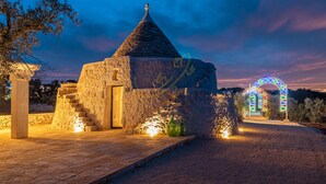 A stone building with a conical roof, a staircase leading to a door, and a colorful illuminated archway in the background. - TD ApulianRetreat Sea view trullo w/heated pool (Costa Merlata)