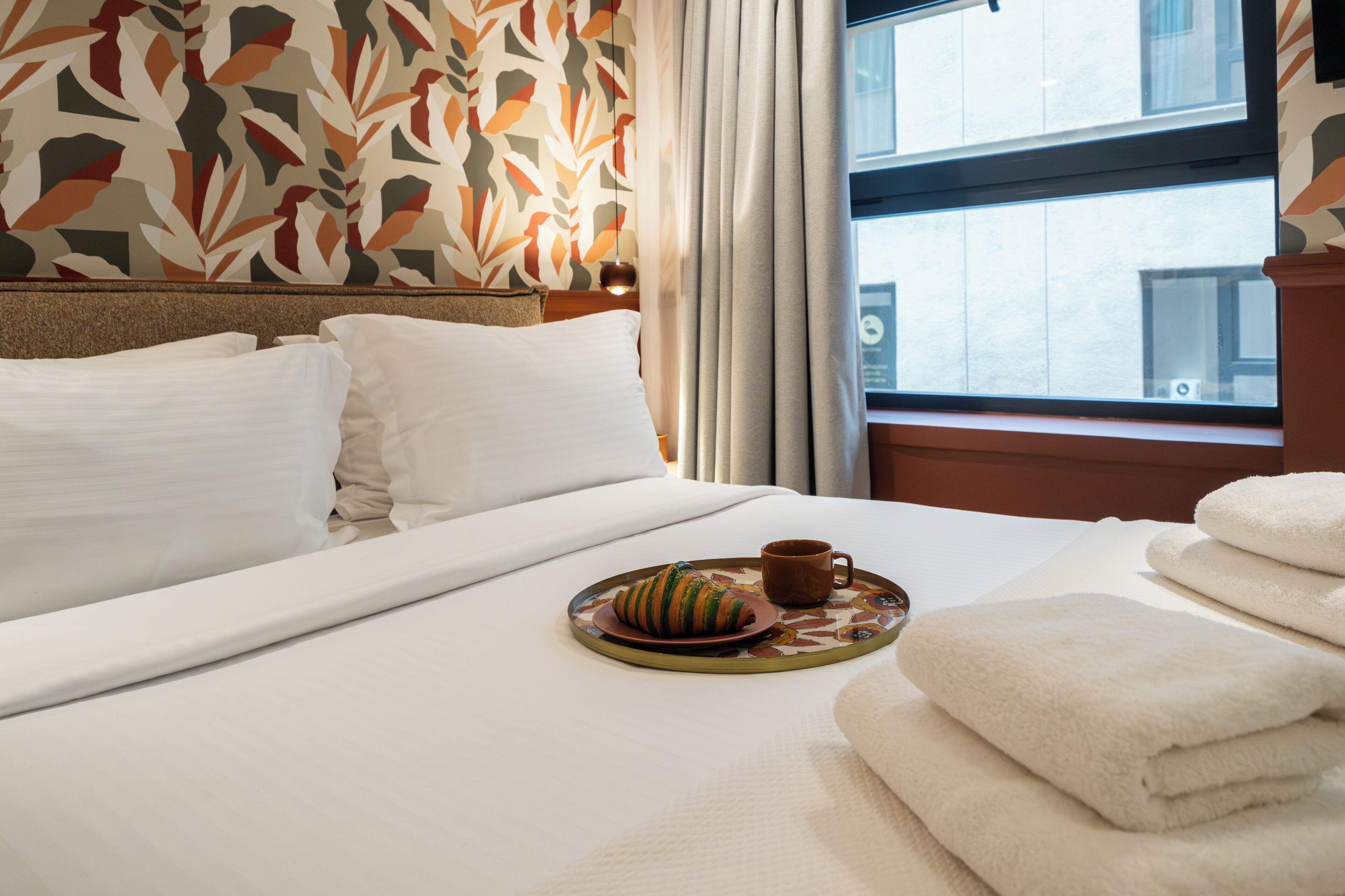 Une chambre d’hôtel avec un lit bien fait, un plateau avec un croissant et une tasse, et une fenêtre avec des rideaux.