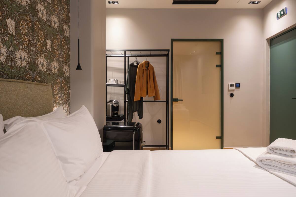 A hotel room with a bed, a desk with a coffee machine, a hanging coat, and a door with an emergency exit sign.