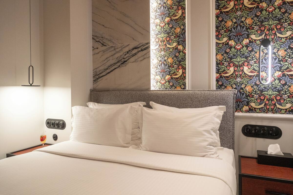 A hotel room with a bed, a nightstand, a lamp, and a window with a floral pattern.