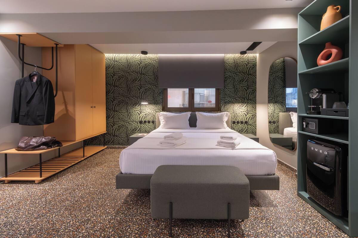 A modern hotel room with a large bed, a bench, a wardrobe, and a minibar.