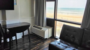 A room with a large window, a flat-screen TV mounted on the wall, a table, a chair, and a leather sofa. - Beachfront Studio fit for a King (Daytona Beach)