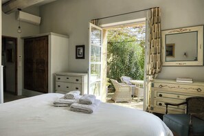 A bedroom with a bed, a dresser, a chair, and an open door leading to a balcony with a view of greenery.