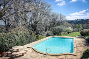 A rectangular swimming pool with a stone deck, surrounded by trees and shrubs.
