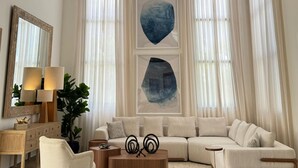 A spacious living room with a large sectional sofa, a coffee table, and a side table. There are two large windows with sheer curtains, a floor lamp, and a mirror on the left wall. - 3-br Villa With Private Pool in Punta Cana Village (Punta Cana)