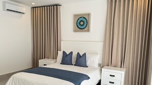 A neatly arranged bedroom with a bed, two bedside tables, a nightstand, a wall-mounted air conditioner, and a decorative wall piece. - 3-br Villa With Private Pool in Punta Cana Village (Punta Cana)