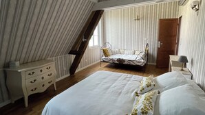A bedroom with a sloped ceiling, a bed with white bedding and gold-patterned pillows, a nightstand, a chair, and a wooden door.
