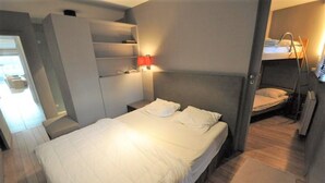 Une chambre d’hôtel compacte comprenant un lit simple, une armoire intégrée, une table de chevet avec une lampe et un lit superposé dans un coin.