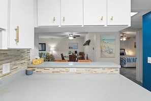 A modern kitchen with white cabinets and gold handles, a dining area with a round table and chairs, and a bedroom visible through an open doorway.