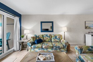Sala de estar com sofá, mesa de centro, mesinha lateral, abajur, espelho e vista através de uma porta de vidro.