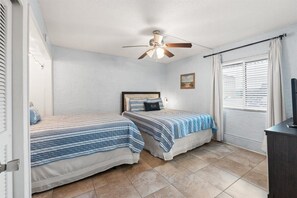 Quarto com duas camas, um ventilador de teto, janela com persianas e uma televisão sobre um suporte.