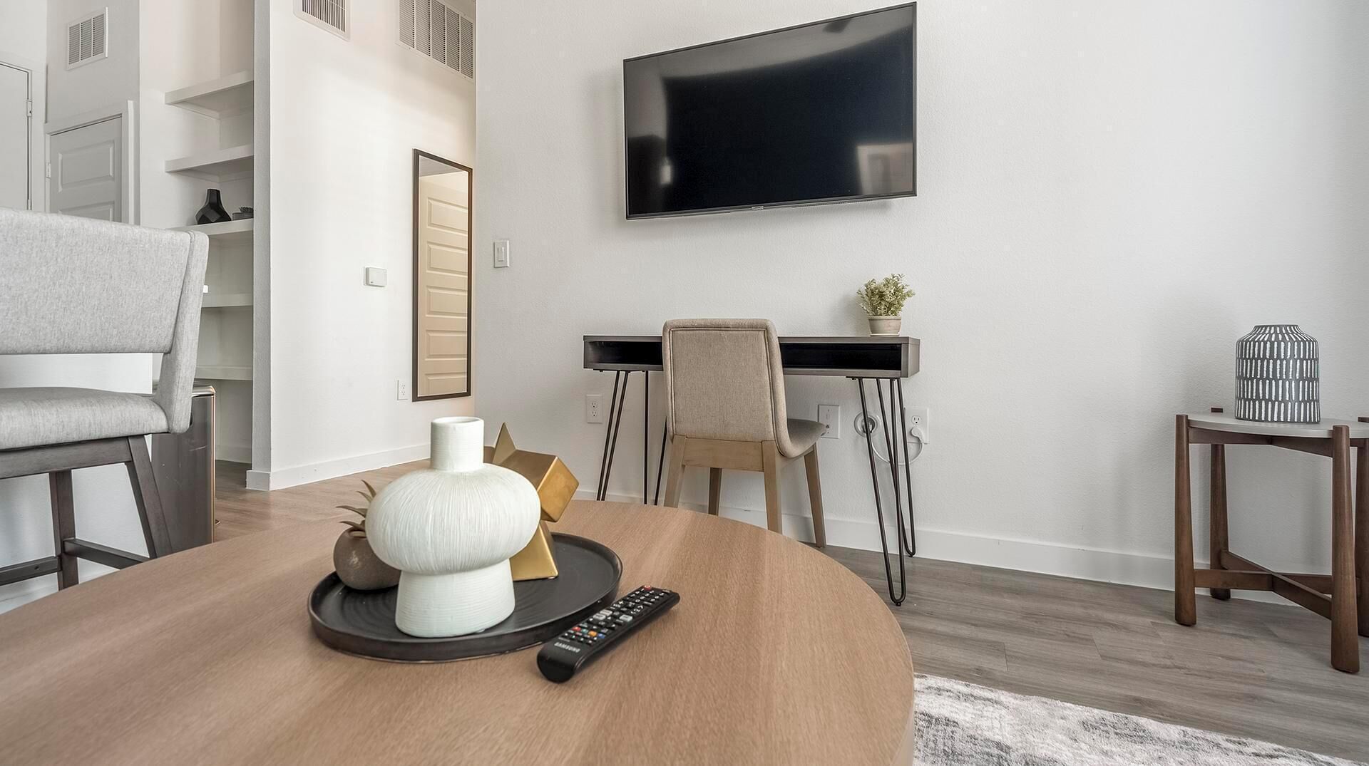 Ein modernes Wohnzimmer mit rundem Couchtisch, einem an der Wand montierten Fernseher, einem Schreibtisch mit Stuhl und einem kleinen Beistelltisch mit Pflanze.