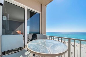 Ein Balkon mit Glastisch und Stühlen, der Blick auf einen Strand mit Menschen und einem klaren Himmel bietet.
