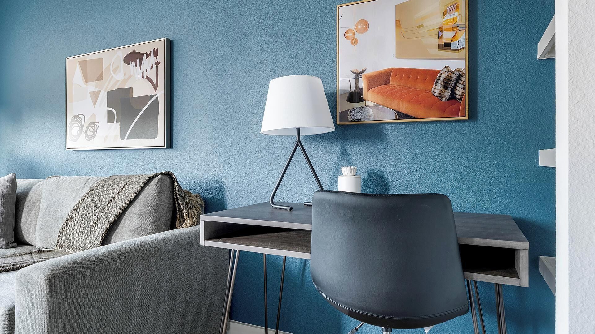Une pièce moderne avec un mur bleu à texture, un canapé gris agrémenté de coussins à motifs, un fauteuil de bureau noir, un bureau avec une lampe blanche et deux œuvres d’art encadrées.