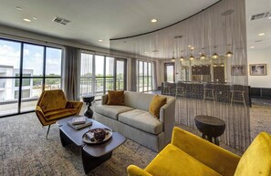 A modern hotel lobby with a grey sofa, yellow armchairs, a coffee table with a book and a glass, and a large window offering a view of the city. - Landing | Beautiful 1BD, Courtyard, Pool (Austin)