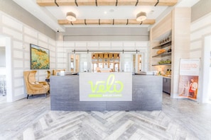 A reception desk with a logo for 'velo shelby farms' in a spacious lobby with a herringbone floor pattern, wooden ceiling beams, and a large painting on the wall.