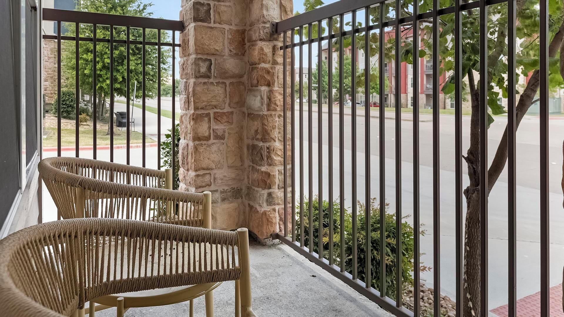 A balcony with two wicker chairs, a stone pillar, and a metal railing overlooking a street.
