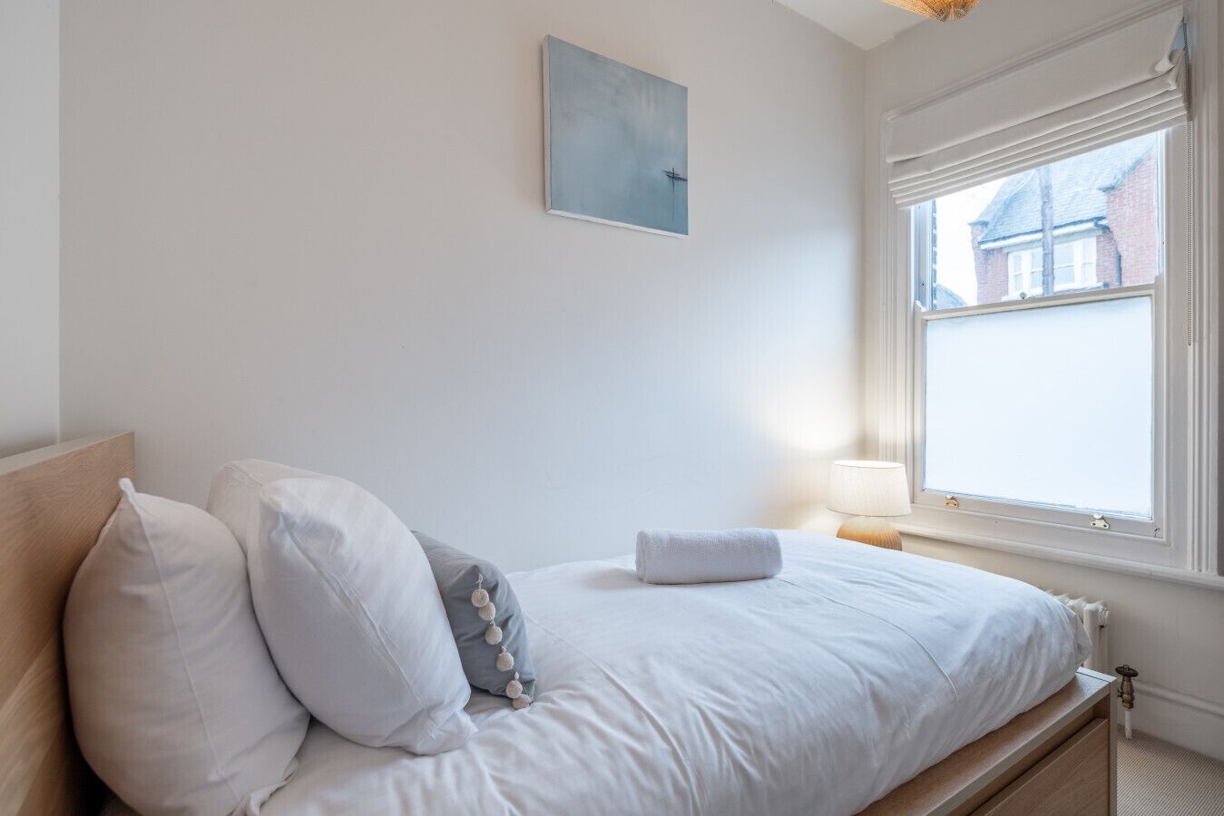 A neatly made bed with white and grey pillows, a small bedside table with a lamp, and a window with blinds.