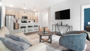 Ein modernes Wohnzimmer mit einer Couch, Sesseln und einem Couchtisch. Im Hintergrund ist eine Küche mit Kühlschrank, Mikrowelle und Barhocker zu sehen.