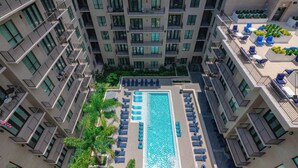 Um hotel moderno com piscina no terraço, rodeado por espreguiçadeiras e palmeiras.