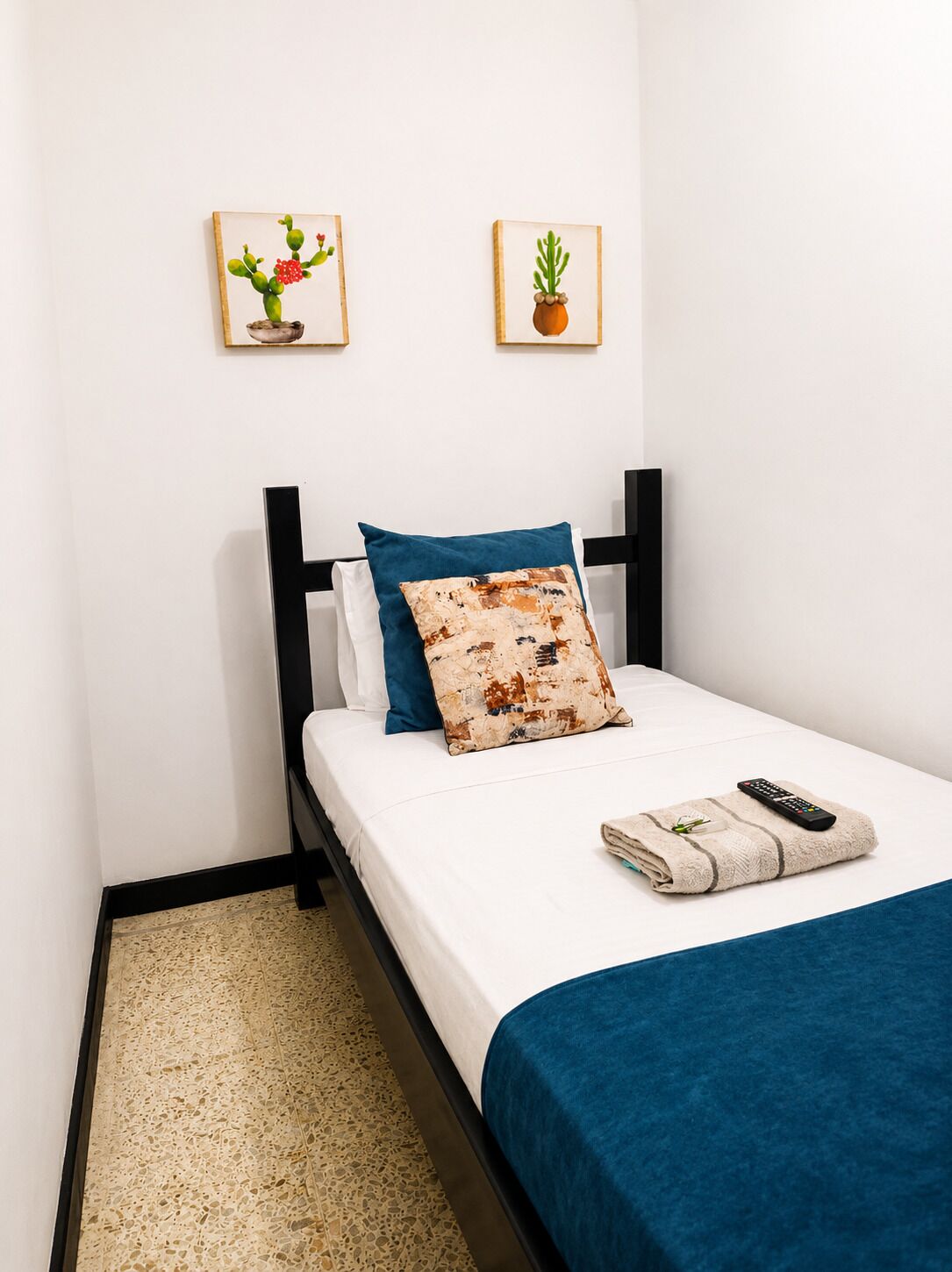 A neatly made bed with a blue blanket, a pillow, and a remote control on the side. There are two framed pictures on the wall above the bed.