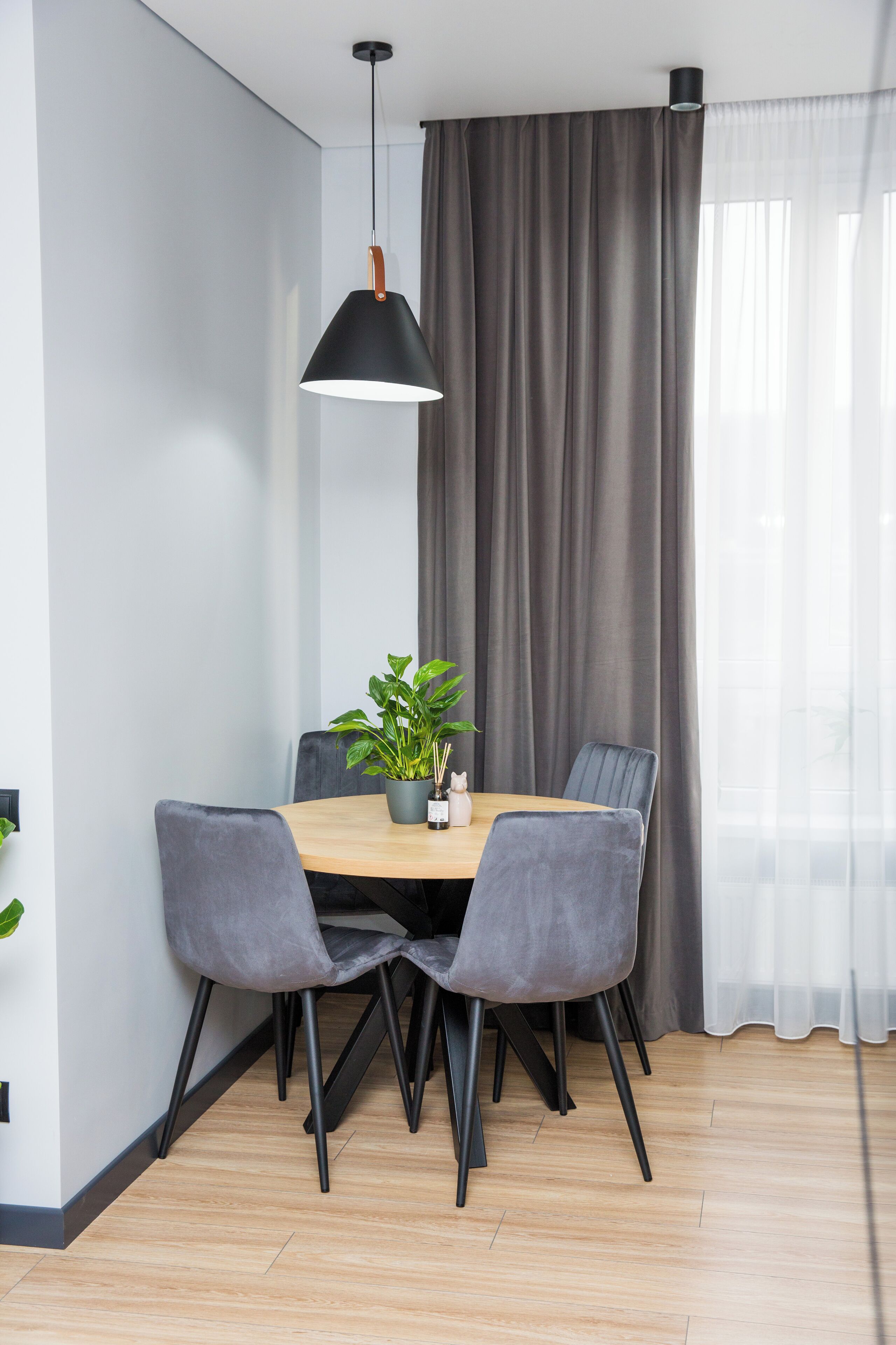 A dining area with a round table, four grey upholstered chairs, a potted plant, and a pendant light.