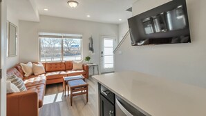 A modern living room with a sectional sofa, a small table, and a kitchen area with a large TV mounted on the wall. - Charming 3-bedroom, 4 Bath Townhome in Arvada, CO (Arvada)