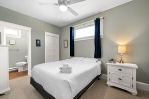 A bedroom with a bed, a nightstand, a ceiling fan, a window with curtains, a door, and a toilet.