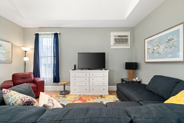 A living room with a grey sofa, a red armchair, a white dresser, a flat-screen TV, and a window with curtains.