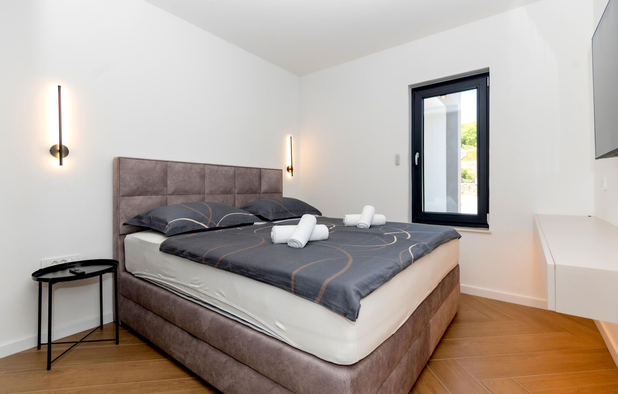 Un dormitorio moderno con una cama grande, dos luces fijadas a la pared, una mesita pequeña y un ventanal con vistas.