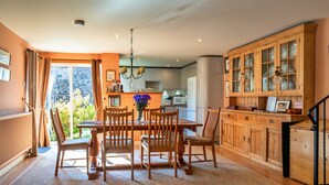 A dining area with a wooden table and chairs, a kitchen with cabinets, and a view of greenery outside. - Cottage in Bovey Tracey Near Dartmoor Park (Newton Abbot)