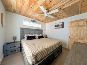 Een slaapkamer met een houten plafond, een bed met geruit beddengoed, een nachtkastje, een bureau en een raam met jaloezieën.