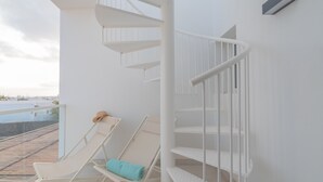 Una scala a chiocciola bianca con ringhiera bianca, una poltrona da relax con asciugamano turchese e vista su spiaggia e mare dal balcone.