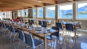 A spacious dining area with multiple tables and chairs, large windows offering scenic views of a lake and mountains, and wooden ceiling beams.
