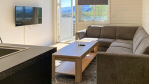 A modern living room with a sofa, a coffee table, a TV mounted on the wall, and a sliding glass door leading to a balcony with a mountain view.
