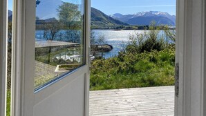 A view through a window of a wooden deck overlooking a river, mountains, and greenery. - 2 bedroom lovely home in ørsta (Ørsta)