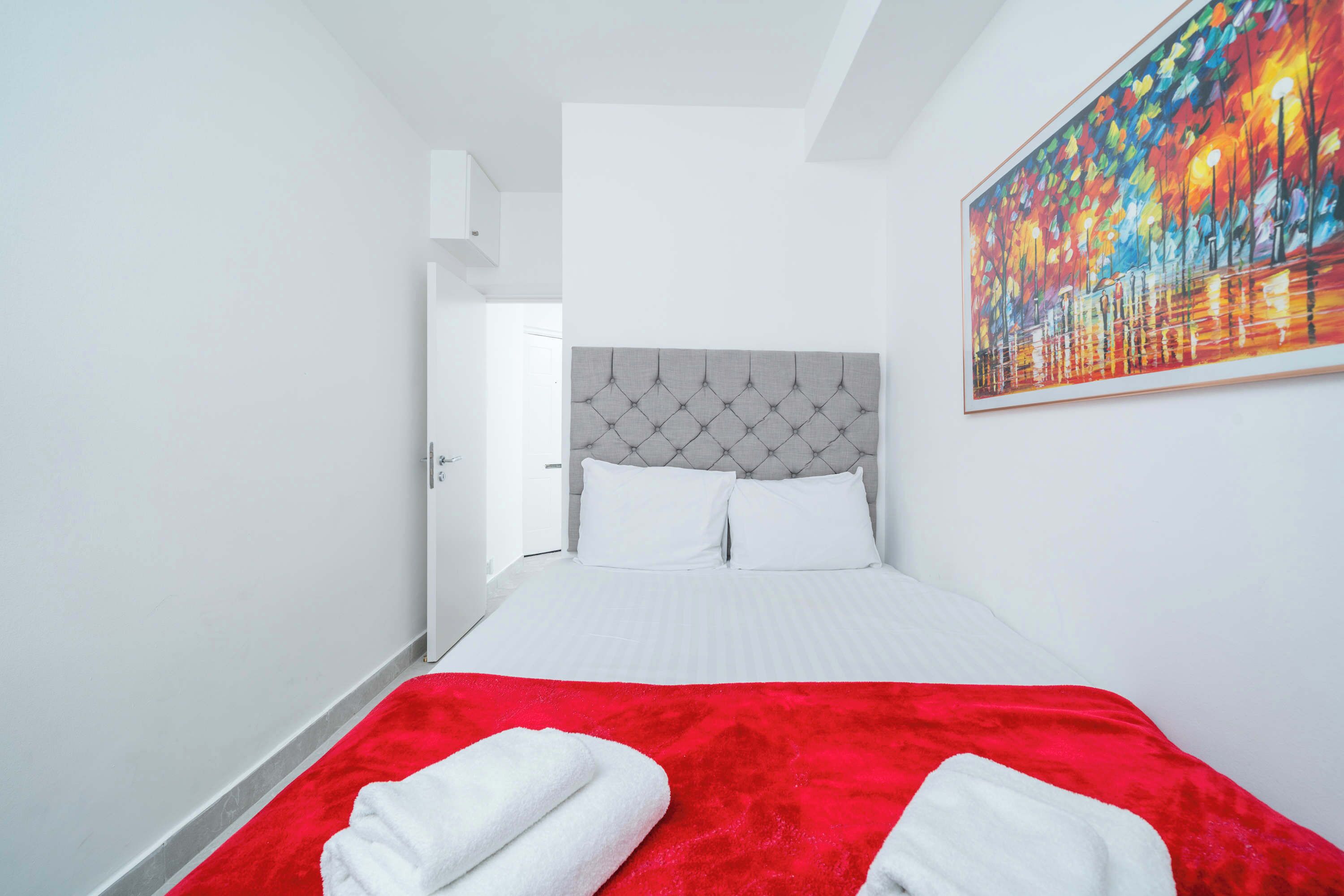 A hotel room with a bed, white bedding, a red blanket, and a painting on the wall.