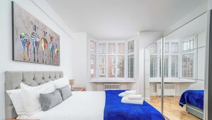 Un dormitorio con cama, espejo, ventana con contraventanas y un cuadro en la pared.