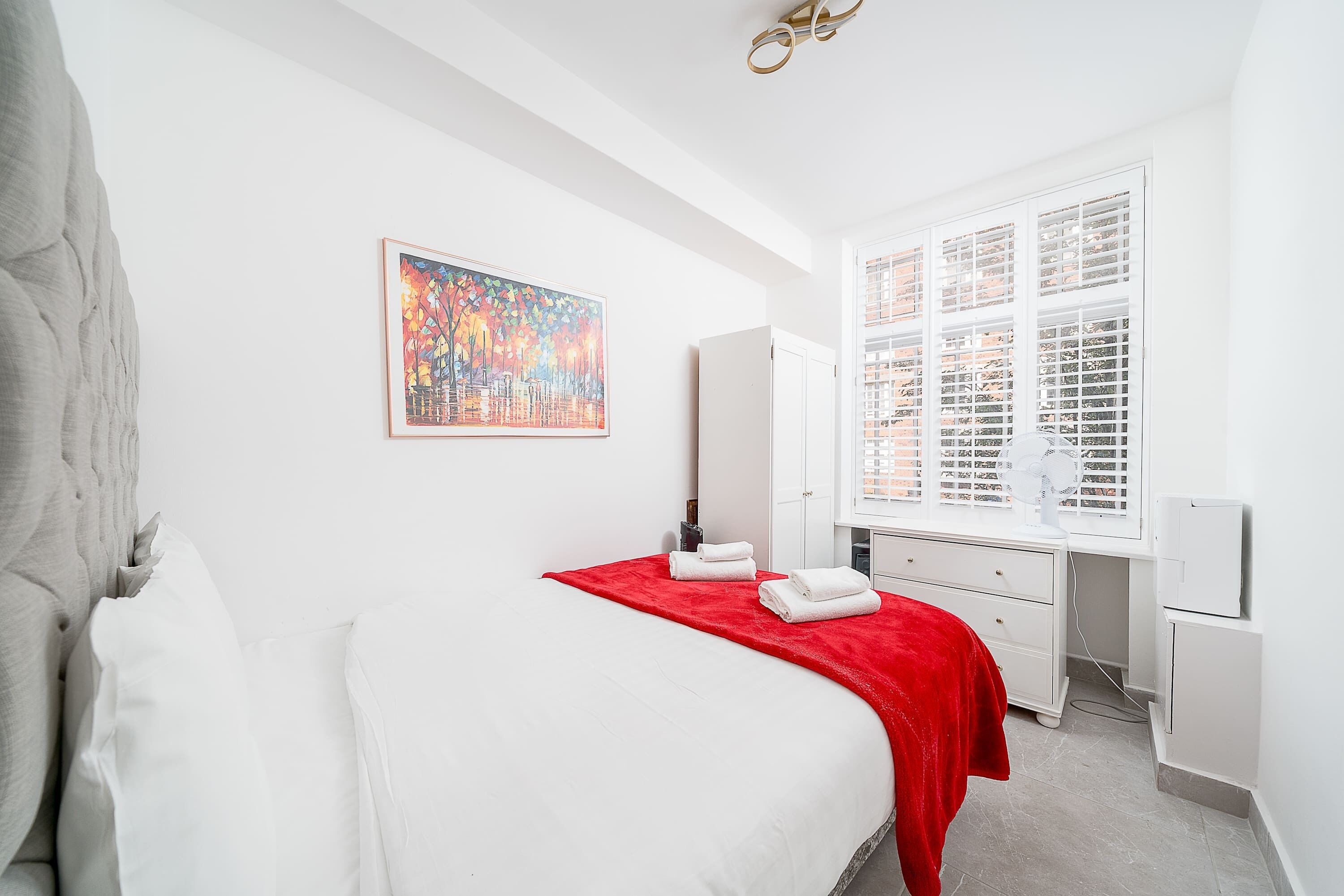 Ein Schlafzimmer mit einem Bett, einer roten Decke, einer weißen Kommode, einem Schreibtisch, einem Fenster mit Jalousien und einem Bild an der Wand.