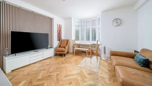 Una sala de estar moderna con un televisor de pantalla plana, un sofá, una mesa de centro de vidrio, piso de madera en patrón de espiga, una zona de comedor con mesa y sillas, y una ventana con persianas.