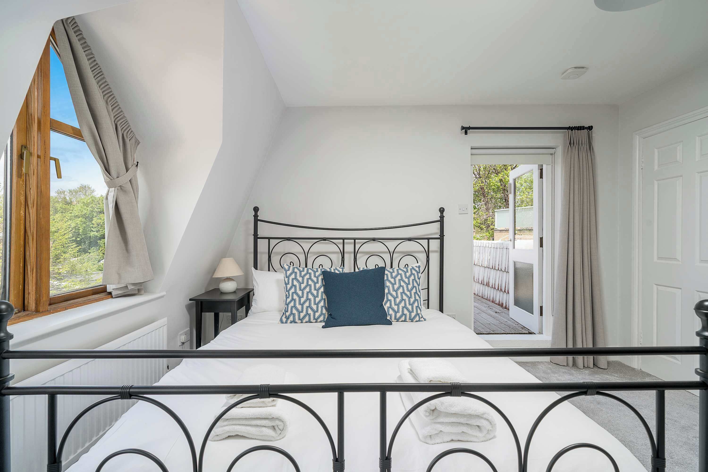 A bedroom with a bed, a nightstand, a window with curtains, and a view of the outdoors.