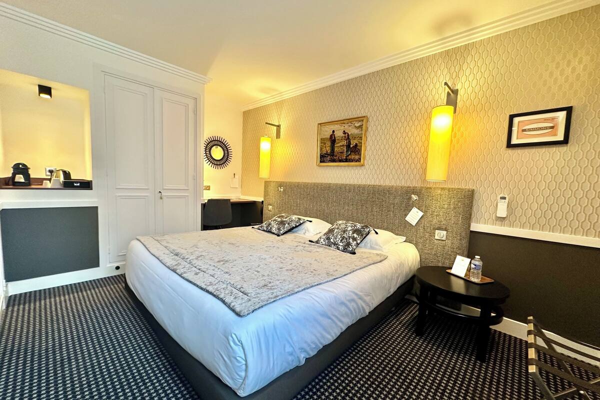 A hotel room with a large bed, a desk, and a chair. There is a mirror, a picture on the wall, and a small table with a water bottle.