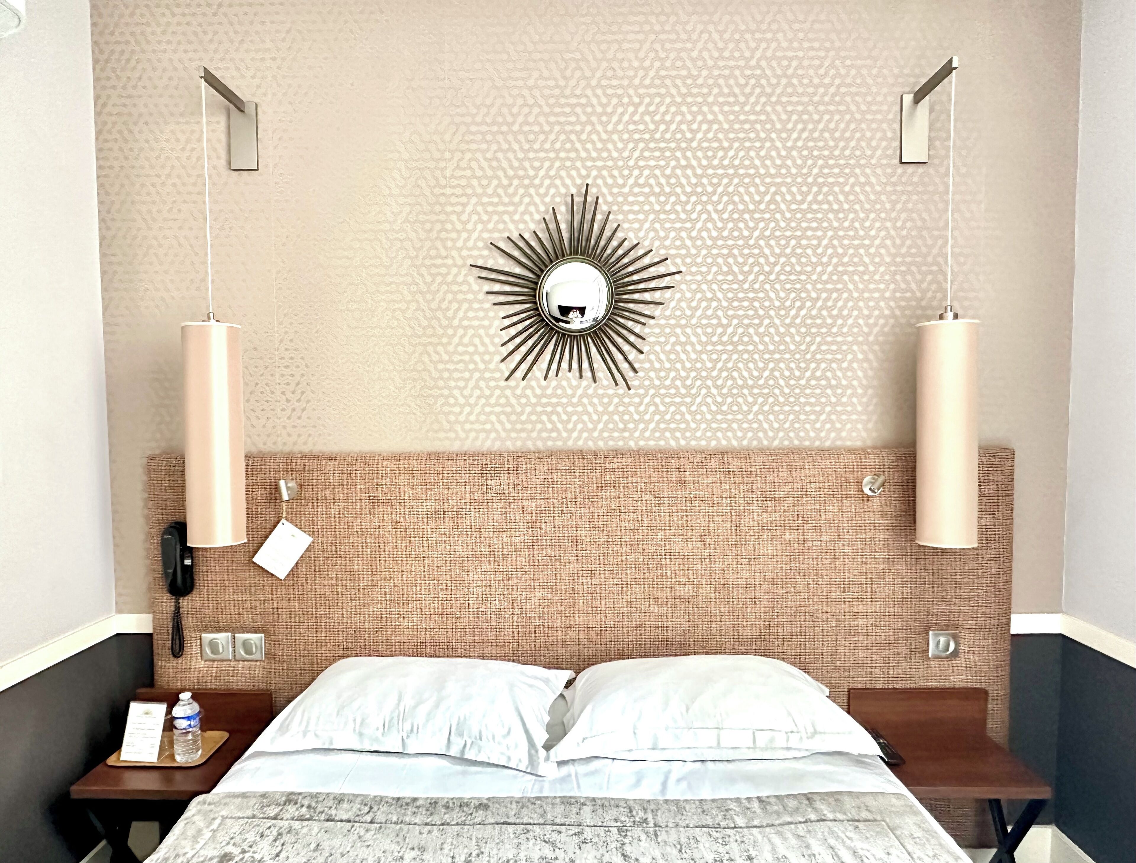 A hotel room with a textured brick wall, a sunburst mirror, two pendant lights, a bed with white linens, a bedside table with a water bottle and a menu, and a telephone.