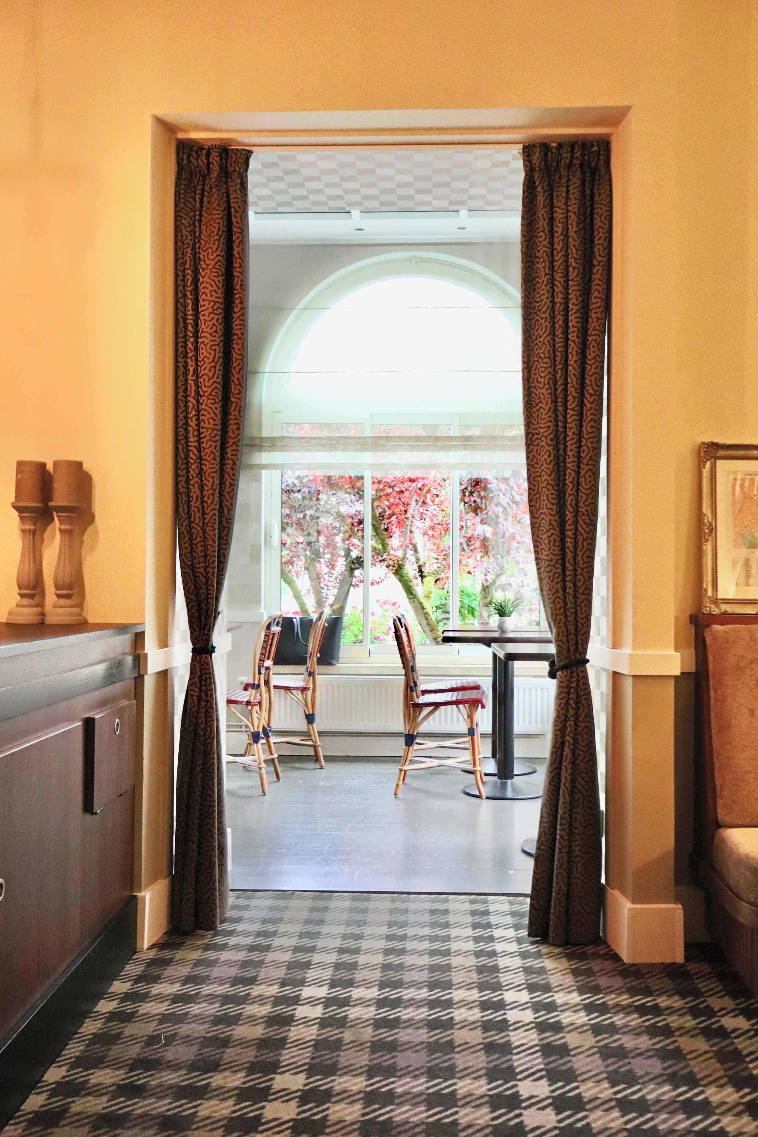 A hallway with patterned carpet, a wooden cabinet, two chairs, a table, and a window with curtains.