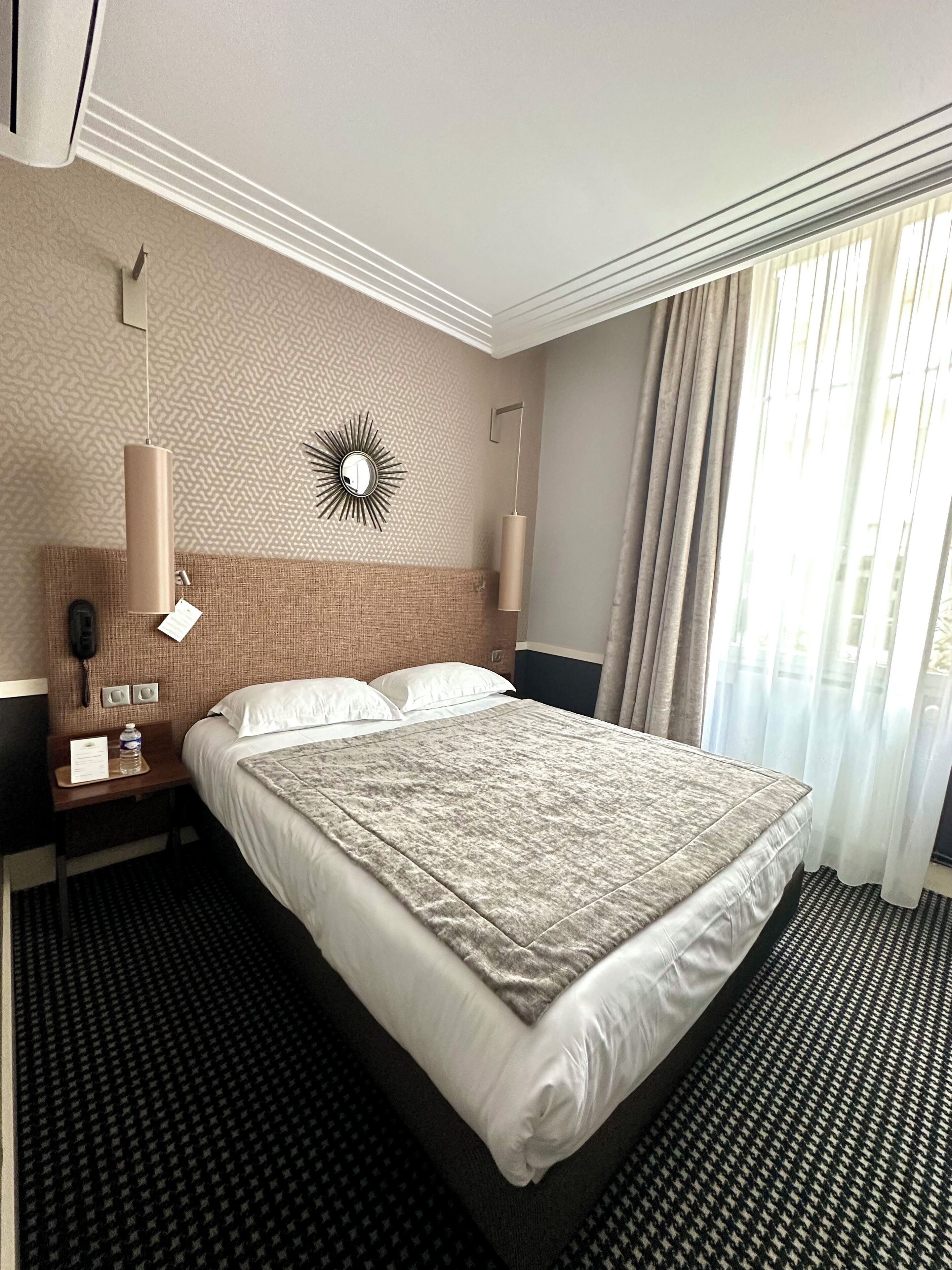 Ett hotellrum med en stor säng, en sänggavel med en spegel, en väggklocka, en telefon och ett fönster med gardiner.