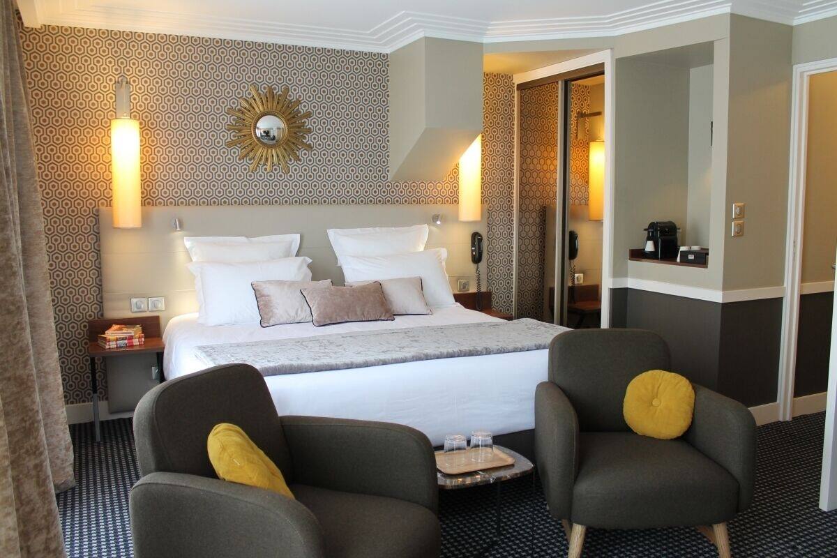 A hotel room with a large bed, two armchairs, a bedside table with a lamp, a mirror, and a patterned wall.